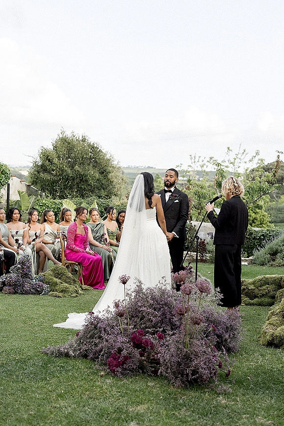Wedding vows as bride in a strapless gown and veil faces groom in tuxedo, with seated bridesmaids and low florals on a garden lawn