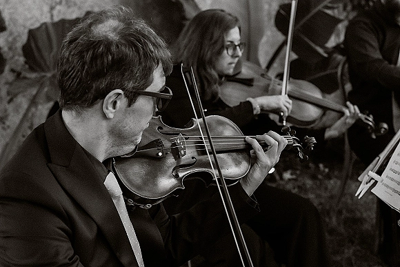 Wedding musicians in suits playing violins beside a music stand with sheet music, performing outdoors against a stone wall