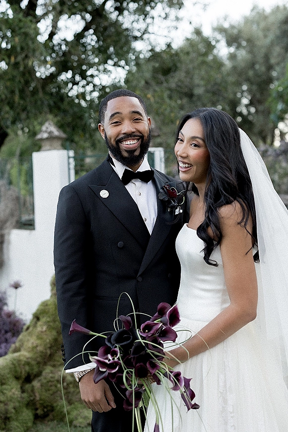 Couple portrait of bride in strapless dress and veil holding a calla lily bouquet beside groom in black tuxedo in a garden setting