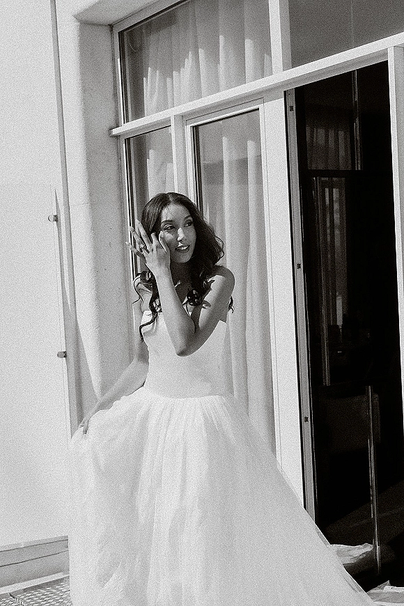 Bridal portrait in black and white of a bride standing by a window, wearing a strapless gown and veil, showing her ring near curtains and balcony railing