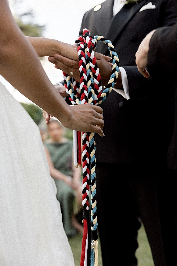Handfasting ceremony with handfasting cords binding bride and groom hands, wedding ring visible, with guests and greenery behind