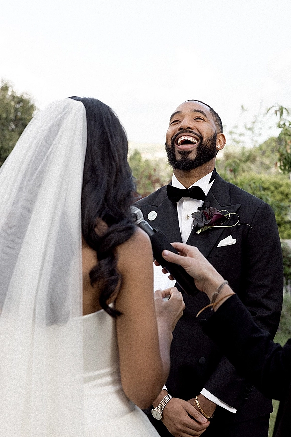 Wedding vows as bride reads vows into a microphone, cathedral veil flowing, while groom in black tuxedo laughs under open sky greenery
