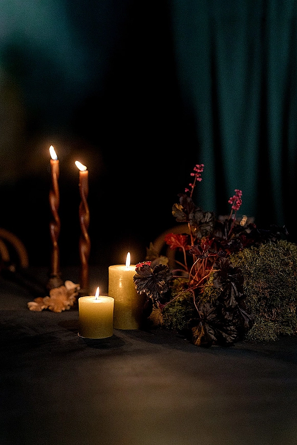 Wedding candle decor with taper and pillar candles in holders, nestled among moss and foliage on table linen against dark drapery background