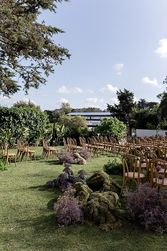 Ceremony setup with wood chairs lining a garden aisle, moss decor and purple flowers on a lawn under blue sky near hedges