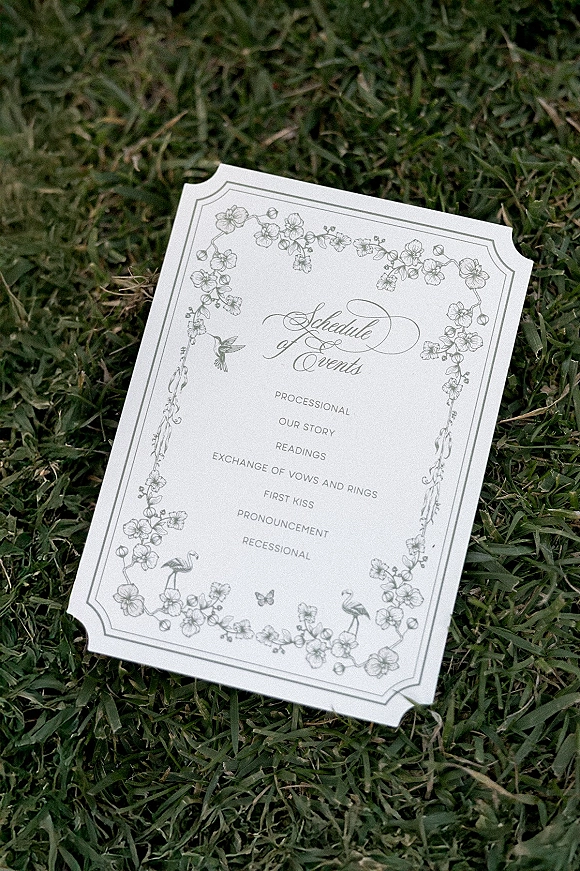 Wedding program card with calligraphy typography and floral border illustration resting on grass lawn, clean black-and-white design
