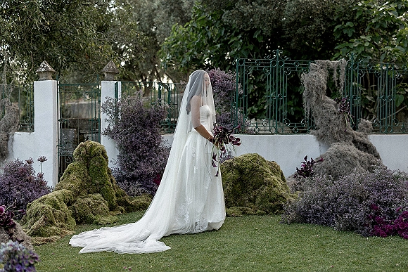 Bridal portrait of a bride in a strapless gown with veil over her face, holding a purple calla lily bouquet on a garden lawn by a wrought iron fence