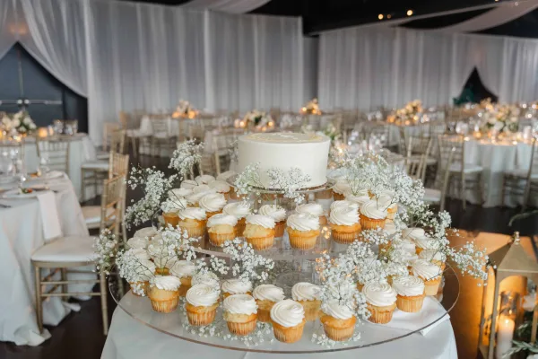 Wedding cupcake display arranged on an acrylic cupcake tower stand, with white draping and round reception tables in the background.
