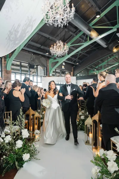 Ceremony recessional, a just married aisle exit with smiling newlyweds amid gold lanterns in a loft venue with brick walls and steel beams.