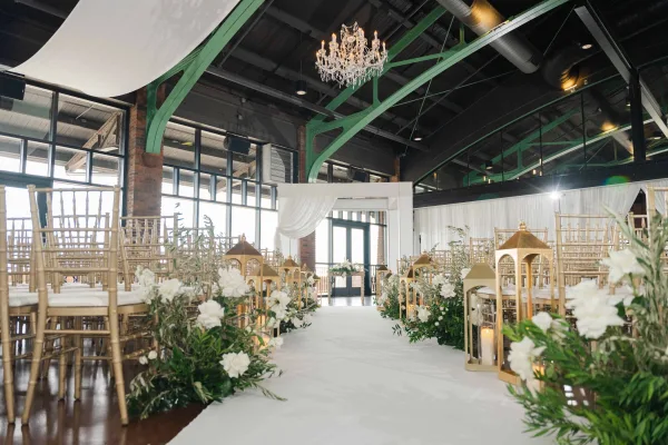 Ceremony aisle design with gold lanterns, elegant indoor ceremony decor set amid industrial beams and brick columns under a high ceiling.