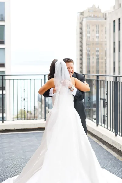 Wedding first look becomes a tender wedding day hug as the veiled bride embraces her partner on a rooftop terrace with a city skyline and water view.