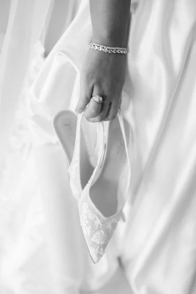 Bridal shoes, delicate lace wedding heels with pointed toes, rest on white fabric beside an engagement ring.