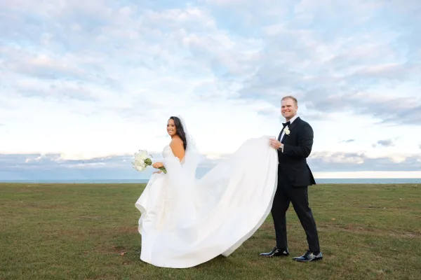 Couple portrait of a bride and groom walking across a grassy field, her bridal bouquet in hand, with a cloudy sky and water horizon in the distance.