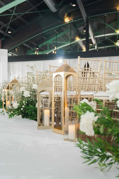 Ceremony aisle decor creates a candlelit ceremony aisle with gold lanterns, set in an indoor venue with white drapery and exposed ceiling beams.