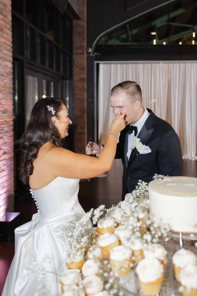 Wedding cake cutting as the bride feeds the groom cake, laughing by a one-tier buttercream cake with baby's breath before a brick wall.