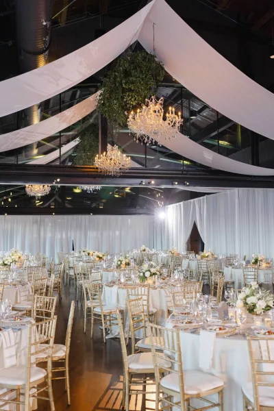 Reception decor for an elegant indoor reception with chandeliers beneath black ceiling beams and white draped walls over polished concrete floor.