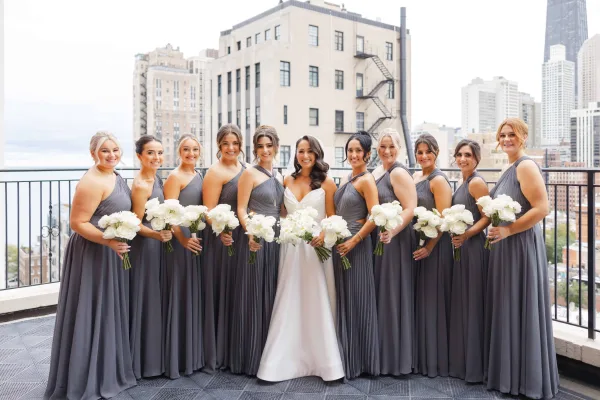Bride and bridesmaids in gray bridesmaid dresses holding white bouquets on a rooftop terrace with a city skyline backdrop.