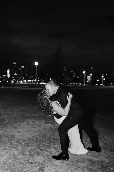 Wedding kiss portrait with the groom dipping the bride in a sleek bridal gown, under city skyline and glowing streetlights on a grass lawn at night.