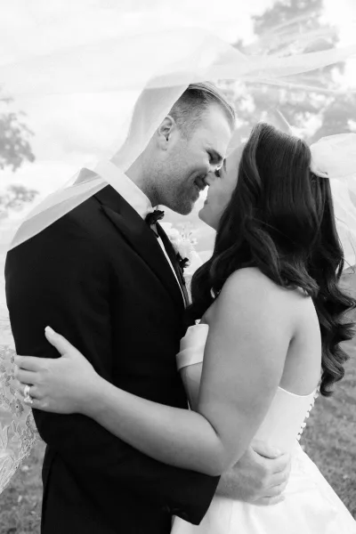 Couple portrait under a veil as the bride and groom lean in for an almost kiss, her engagement ring catching the light on her hand.