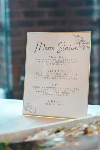 Wedding menu sign used as a food station sign, featuring a lemon illustration, displayed on table linen against a brick wall backdrop.