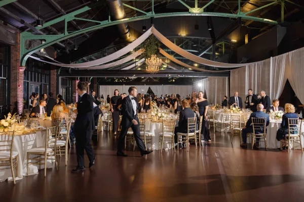 Reception decor with a ceiling draping installation and a chandelier, set in an industrial venue with brick walls and exposed beams.