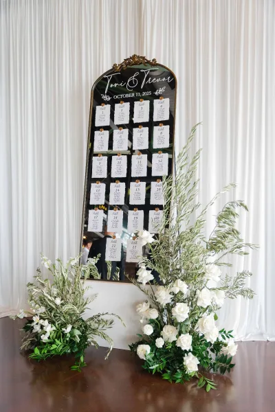 Wedding seating chart presented as a mirror seating chart before soft white drapery on a wood floor.