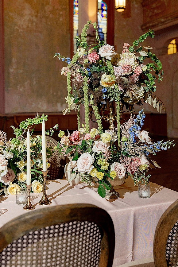 Reception centerpiece featuring a wedding floral centerpiece in a compote bowl with hanging amaranthus, roses, and brass taper candles by stained glass windows