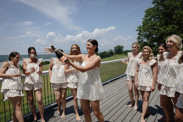 Bridesmaid champagne pop as bridal party in matching pajamas and bride in white slip cheer under champagne spray on lake-view deck