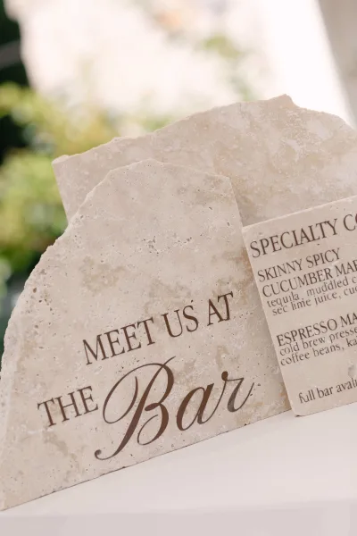 Wedding bar sign with calligraphy lettering on stone signage, paired with a printed cocktail menu card on a white tabletop, greenery blurred behind