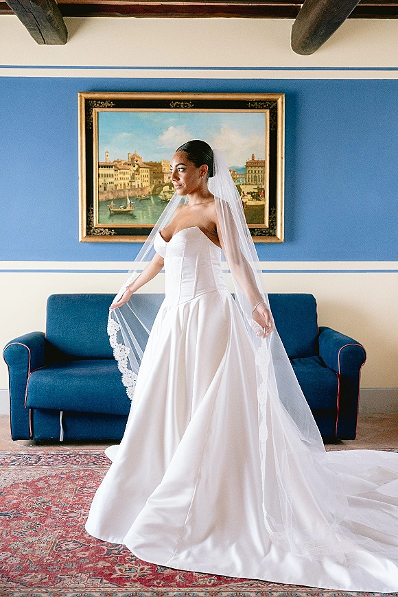Bridal portrait of a bride in a strapless wedding dress with lace-trim veil and jewelry, standing in a blue room with sofa and art