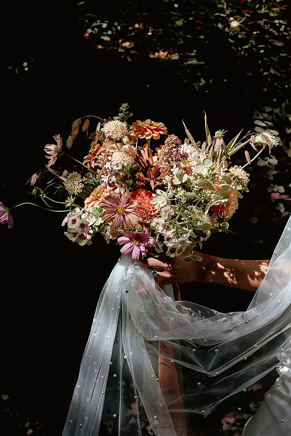 Bridal bouquet of colorful wildflowers with peach and rust blooms, held beside a pearl veil and ring against dappled garden greenery