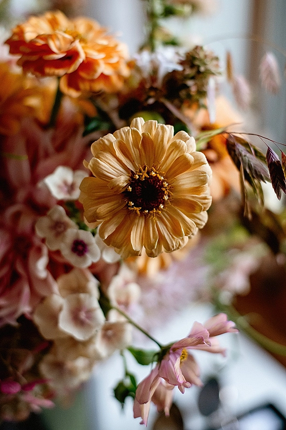 Wedding bouquet of warm-toned wildflowers with dahlias, zinnias, cosmos, and dried grass, hand-tied against a softly blurred indoor backdrop