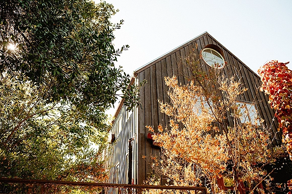 Barn wedding venue with rustic barn exterior and warm string lights, featuring a wooden balcony and round window amid sunlit trees