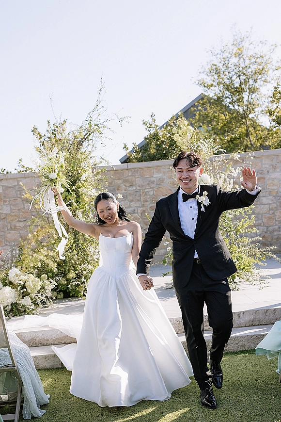 Wedding recessional as bride and groom walk the aisle holding hands, bride raising bouquet with ribbons beside a stone wall and trees