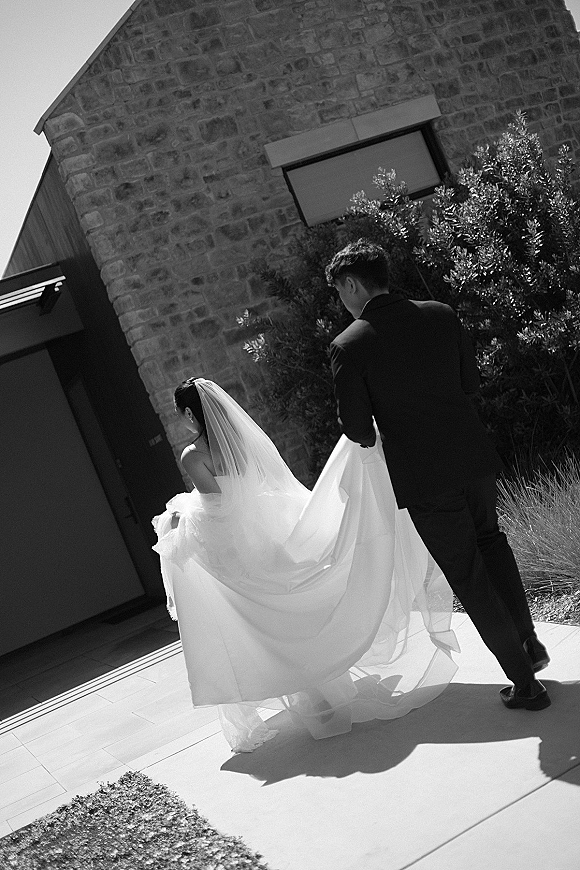 Wedding couple portrait in black and white as bride and groom walk away, groom lifting her dress train by a stone entry walkway