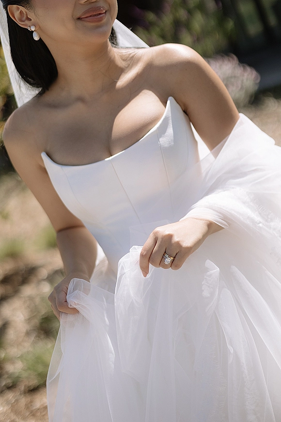 Bridal portrait in a strapless wedding dress, veil draped over her arms and pearl drop earrings, standing on a sunlit garden path