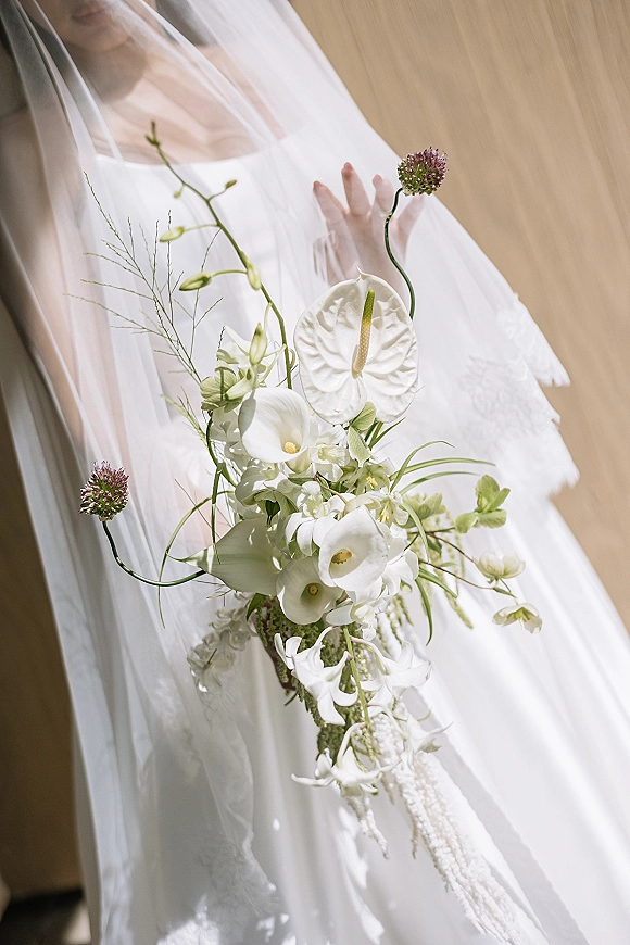 Bridal bouquet with a white calla lily bouquet featuring anthurium, orchids, and greenery, held against a white veil in window light