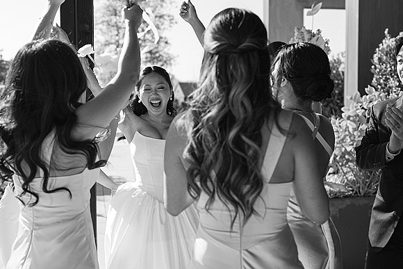 Bridal party moment as bride with bridesmaids cheer in window light, her strapless gown glowing in an indoor reception space with greenery