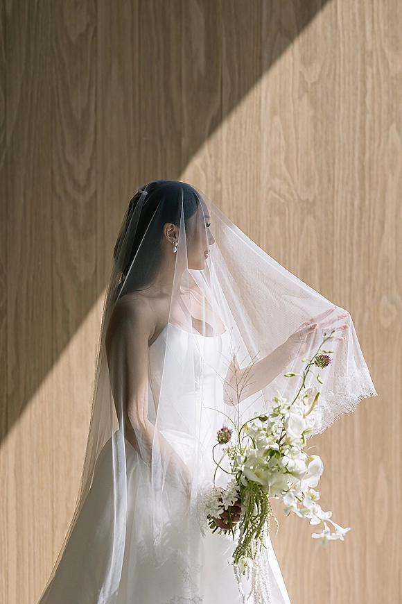 Bridal portrait of a bride with veil in a strapless gown, holding a white orchid bouquet by a wood panel wall in window light