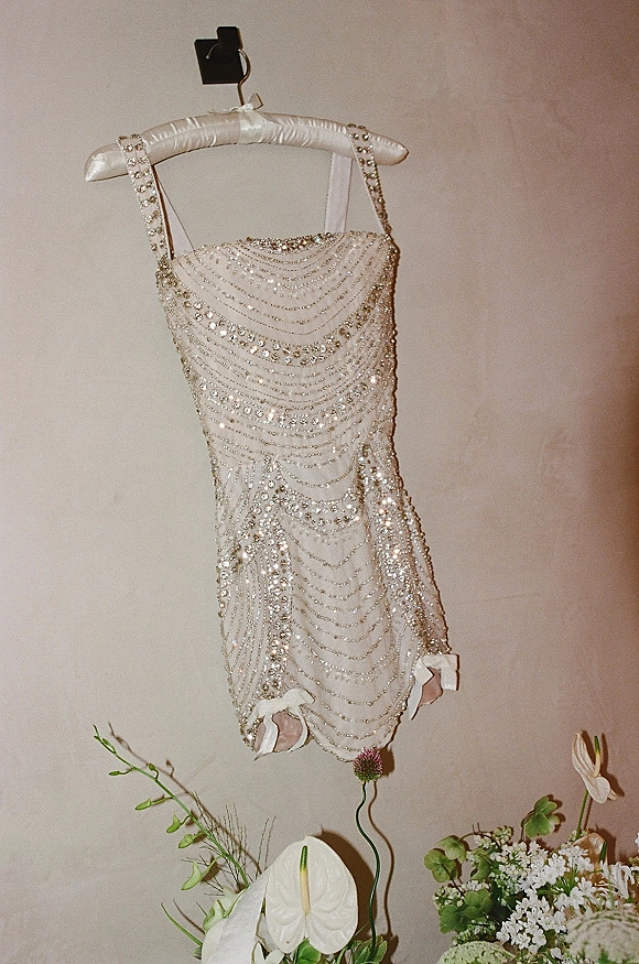 Wedding dress with beaded wedding dress detailing and rhinestone straps on a hanger, finished with ribbon bows and flowers against a neutral wall
