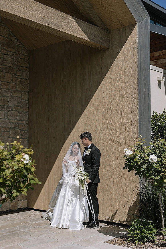 Couple portrait of bride in a cathedral veil and satin dress holding a white bouquet beside groom in black tux under wood overhang