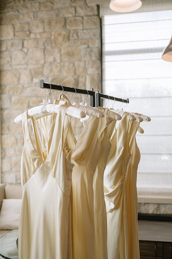 Bridesmaid dresses in champagne bridesmaid dresses tones hanging on a rack, satin sheen, against a stone wall with pendant lights