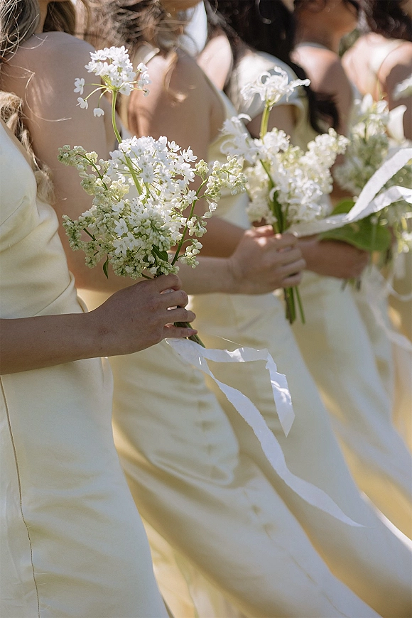 Bridesmaid bouquets of white wildflowers with ribbon streamers held against satin bridesmaid dresses in sunlit outdoor setting