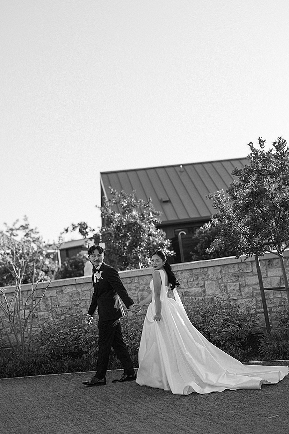 Couple portrait of bride and groom walking away holding hands, looking back, her long veil and dress train by a stone wall outdoors.