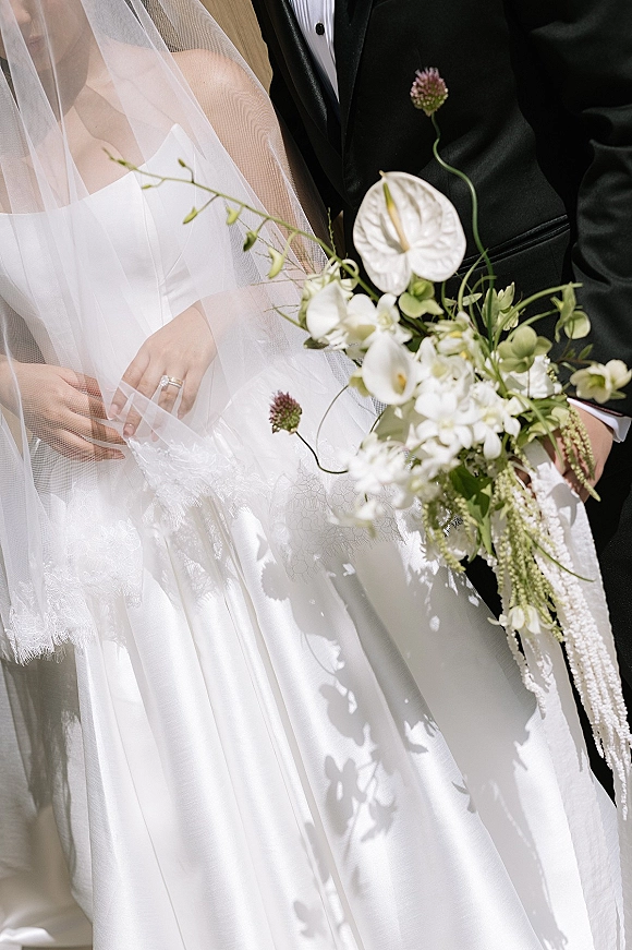 Wedding couple portrait of bride and groom close up, her veil and strapless dress beside a cascading anthurium-orchid bouquet against a neutral wall