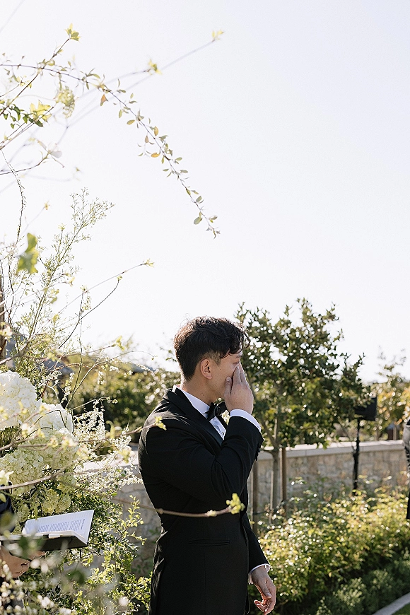 Groom emotional moment as he wipes tears in a black tuxedo and bow tie beside hydrangea florals at an outdoor garden ceremony