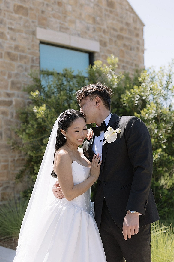 Couple portrait of the bride and groom hug as he kisses her forehead, her long veil and pearl earrings framed by a stone building window.