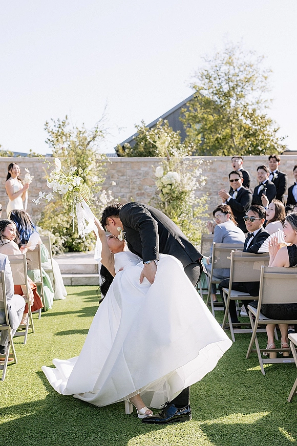 Wedding kiss moment as groom dips bride in the aisle, her bouquet raised overhead, guests cheering beside folding chairs on a sunny lawn