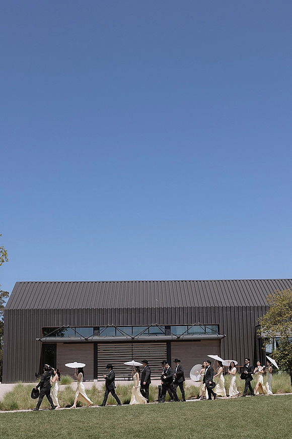Wedding party walk with bridesmaids and groomsmen in black suits and champagne dresses, one carrying a white parasol outside a modern venue