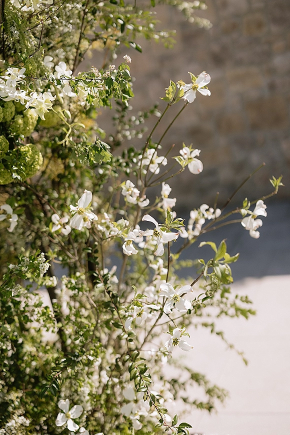 Wedding florals and ceremony flower arrangement with white blossoms and greenery branches on a sunlit outdoor patio by a stone wall