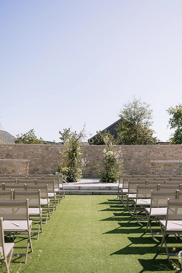 Ceremony setup for an outdoor wedding ceremony with chair rows facing white and green floral pillars on stone steps by a stone wall